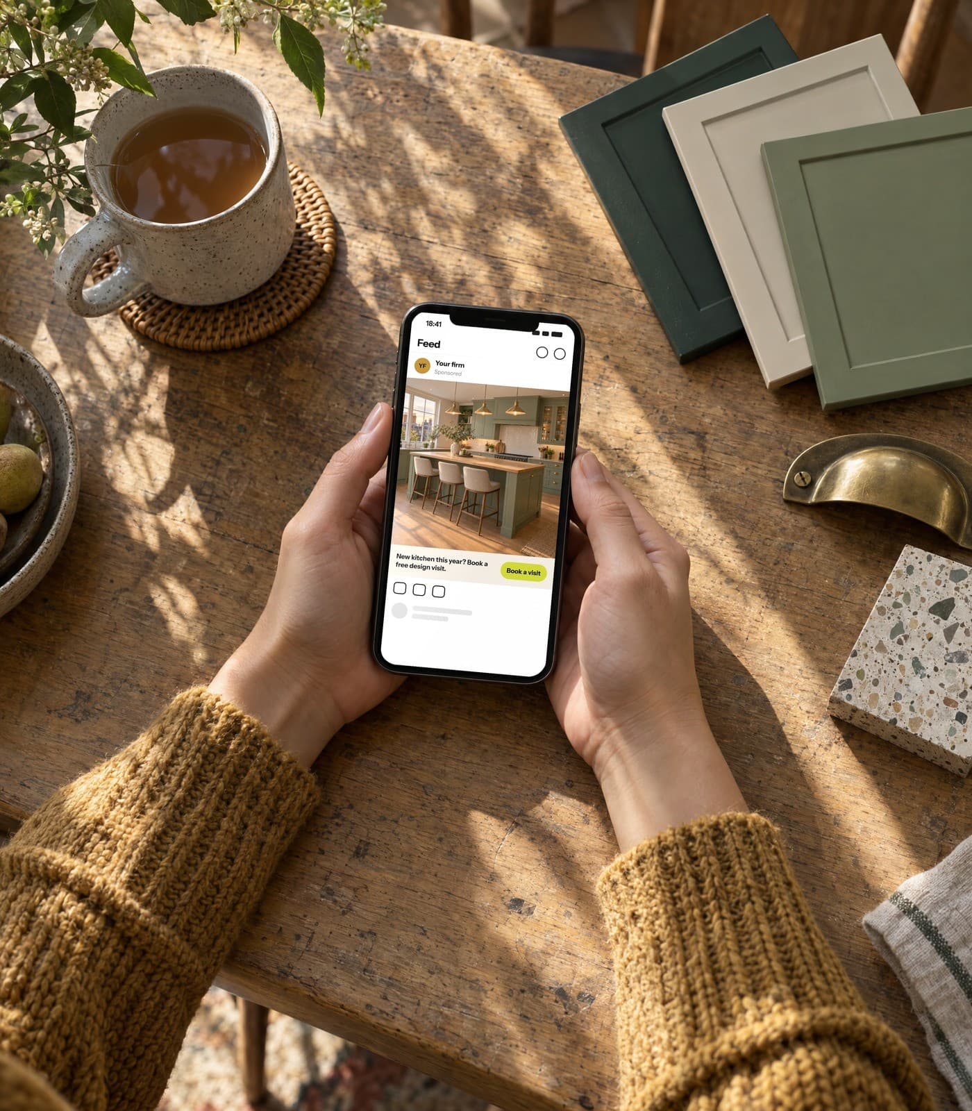 Hands holding a phone over an oak table with kitchen door samples; the phone shows a demo Facebook ad for a kitchen firm.